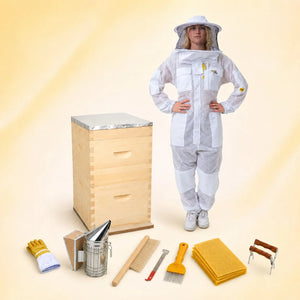 Beekeeper in a ventilated white suit beside a wooden beehive with a full kit of smoker, tools, brush and wax foundation