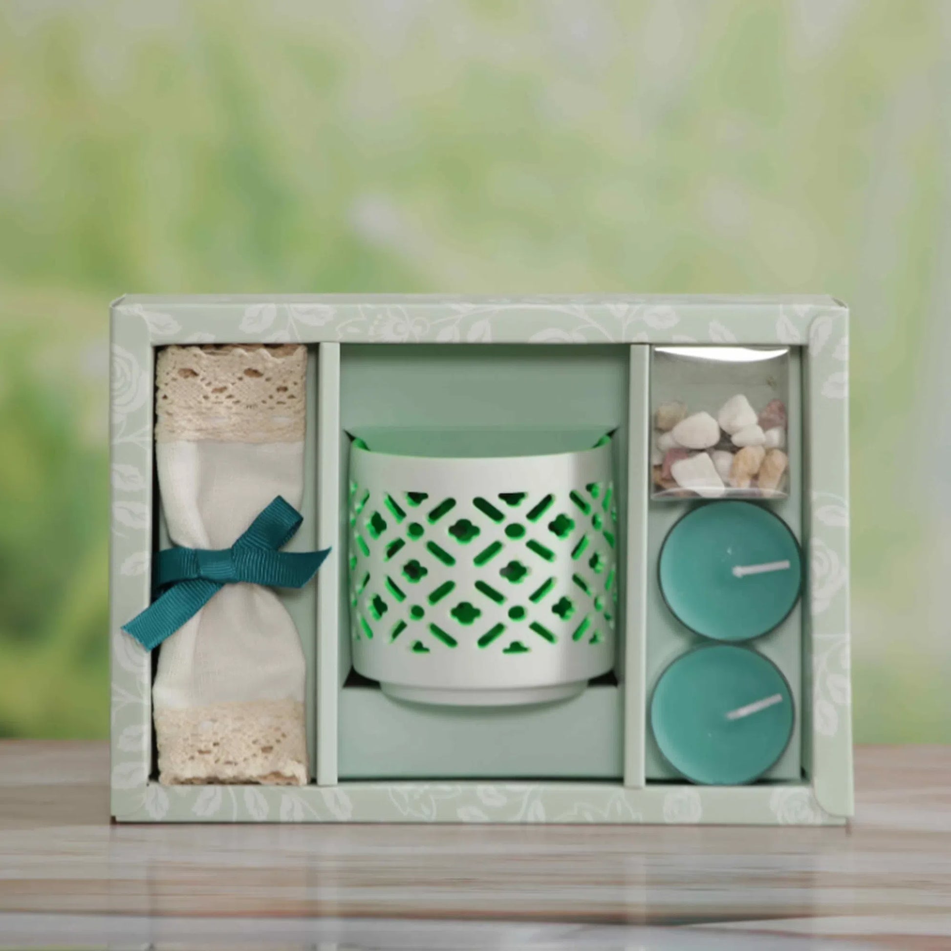 Jasmine Scented Candle, A gift set in a square box with a pale green floral pattern, containing a mint green patterned candle holder, a folded cream fabric item tied with a teal ribbon, and two round teal candles in a small clear container, set on a wooden surface with a blurred green background.