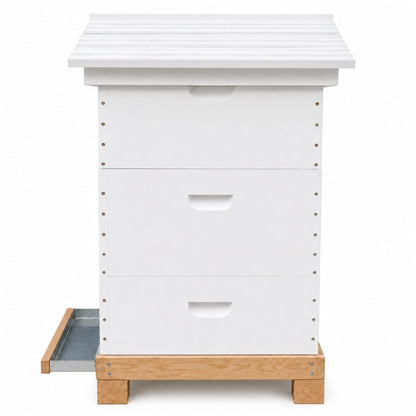 White backyard beehive with a decorative peaked roof and round entrance hole, featuring three dovetailed boxes on a cedar base