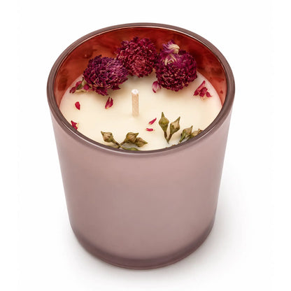 A single-wick candle in a translucent mauve glass jar filled with white wax, decorated with dried rose petals and green leaves on top, set against a plain white background.